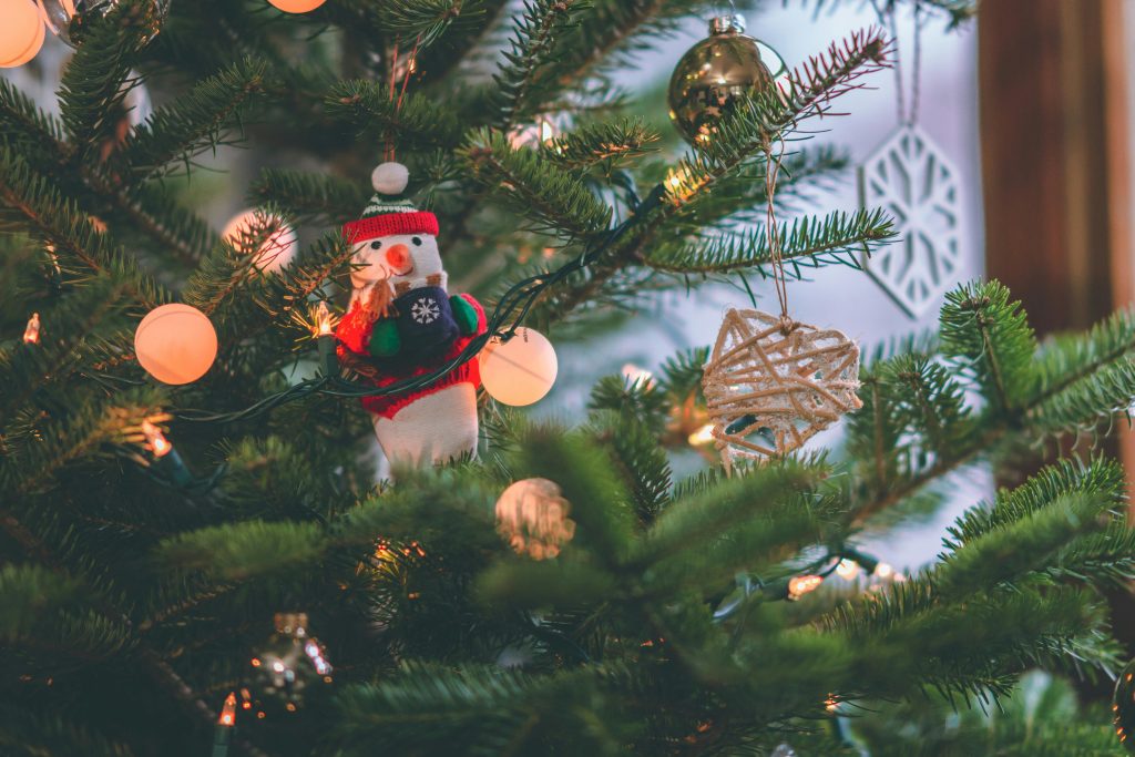 A little snowman ornament hangs on a Christmas tree with bauble lights.