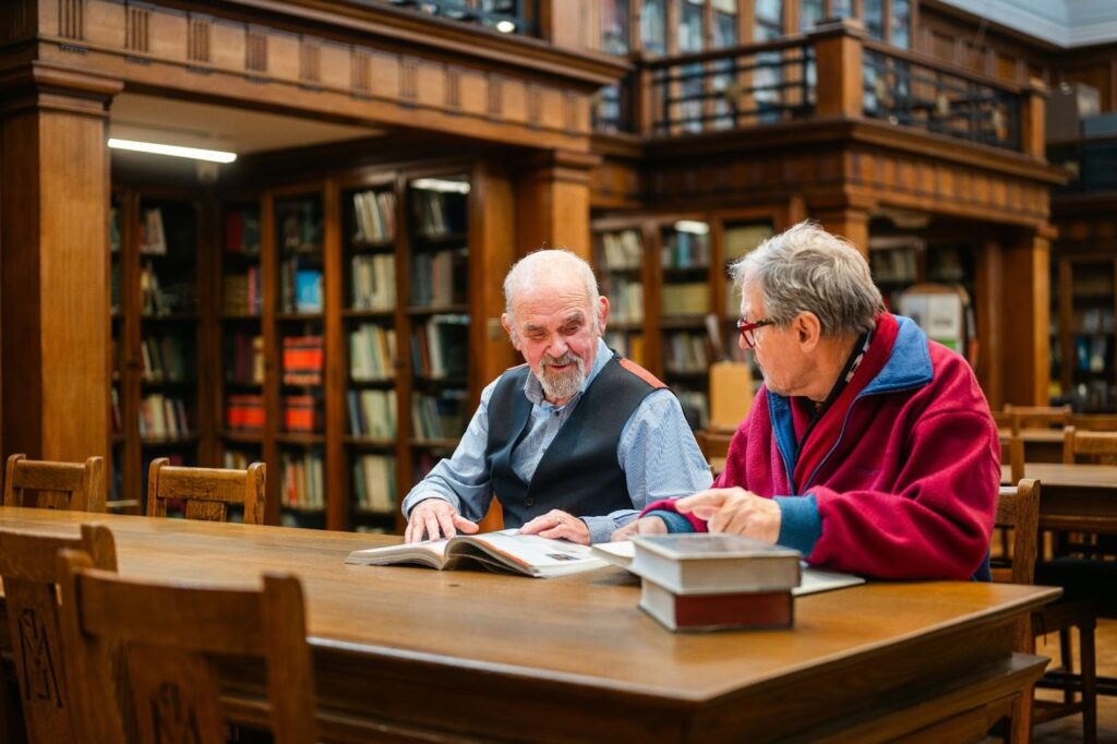Two older men sitting at a wooden table in a library, reading and discussing a book surrounded by bookshelves.