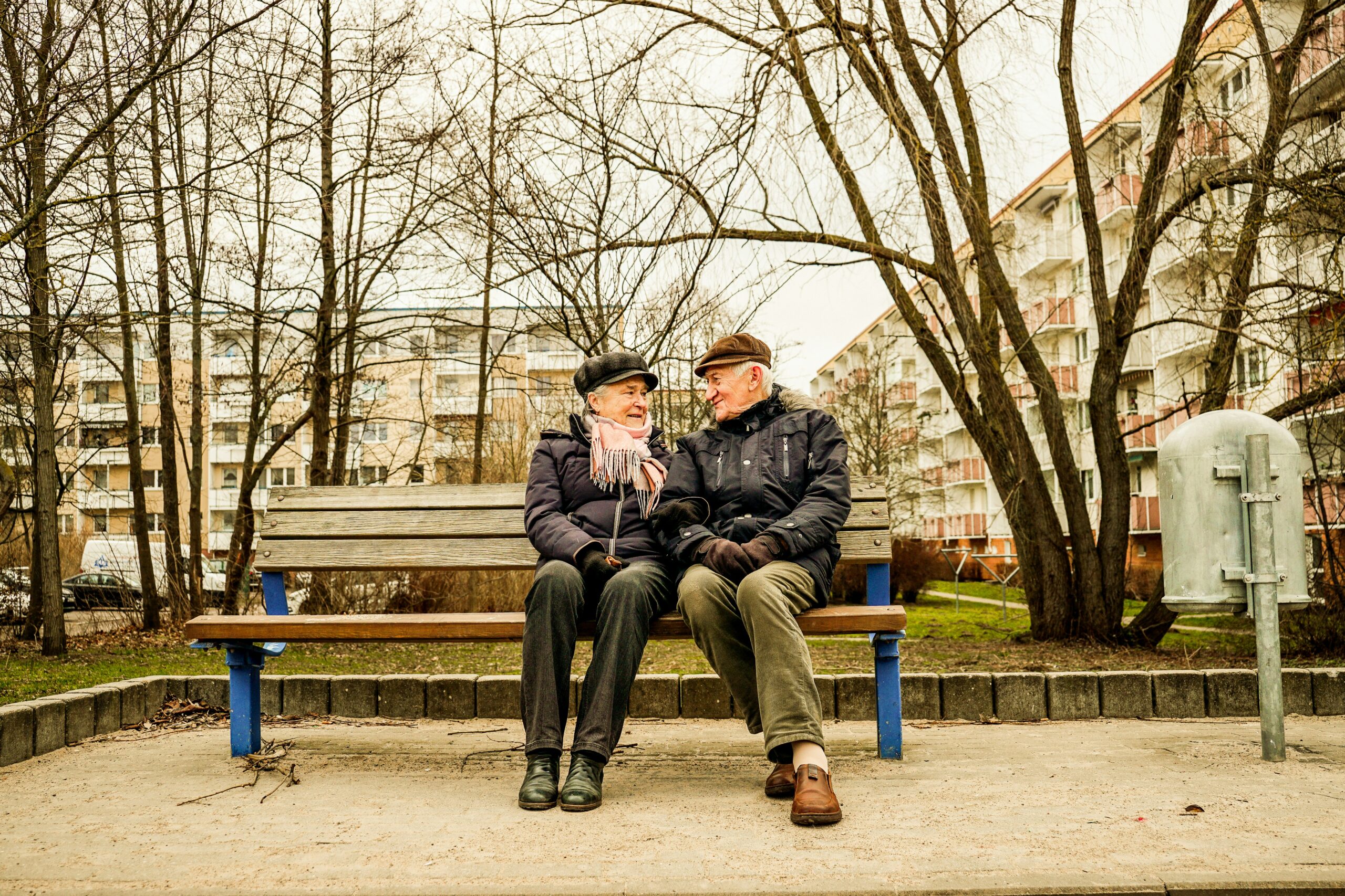 Elderly couple sitting close together on a park bench, smiling at each other on an overcast day with apartment buildings in the background.